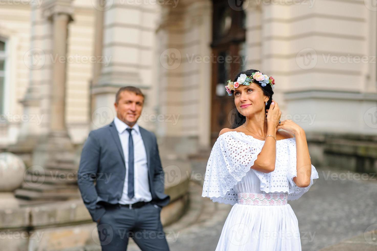 Wedding photo session on the background of the old building. The groom watches his bride posing. Rustic or boho wedding photography.