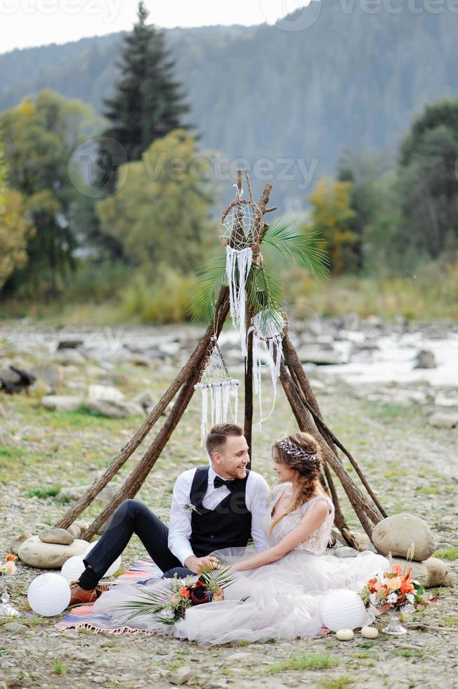 atractiva pareja de recién casados, momento feliz y alegre. el hombre y la mujer con ropa festiva se sientan en las piedras cerca de la decoración de la boda al estilo boho. ceremonia al aire libre. foto