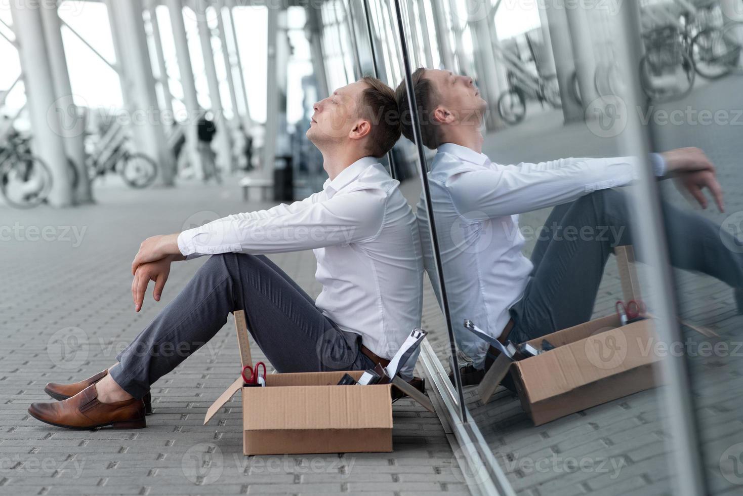 The fired office worker fell to his knees and covered his face due to stress. In front of him is a cardboard box with stationery. The man is unhappy due to a reduction in the robot. photo