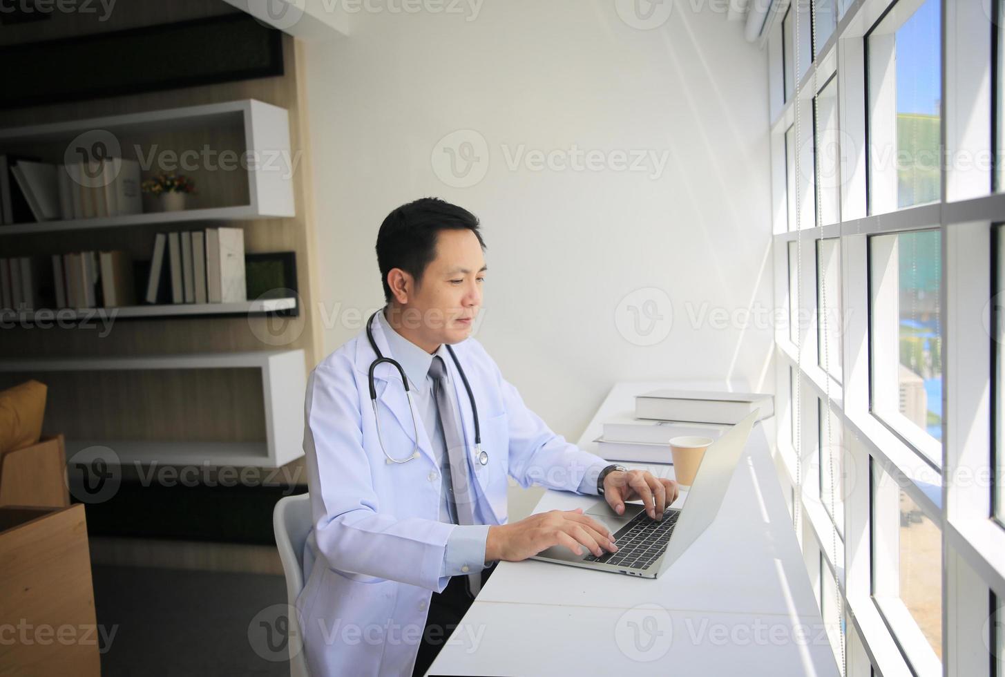 médico masculino asiático usando una computadora portátil junto a la ventana en su oficina. foto