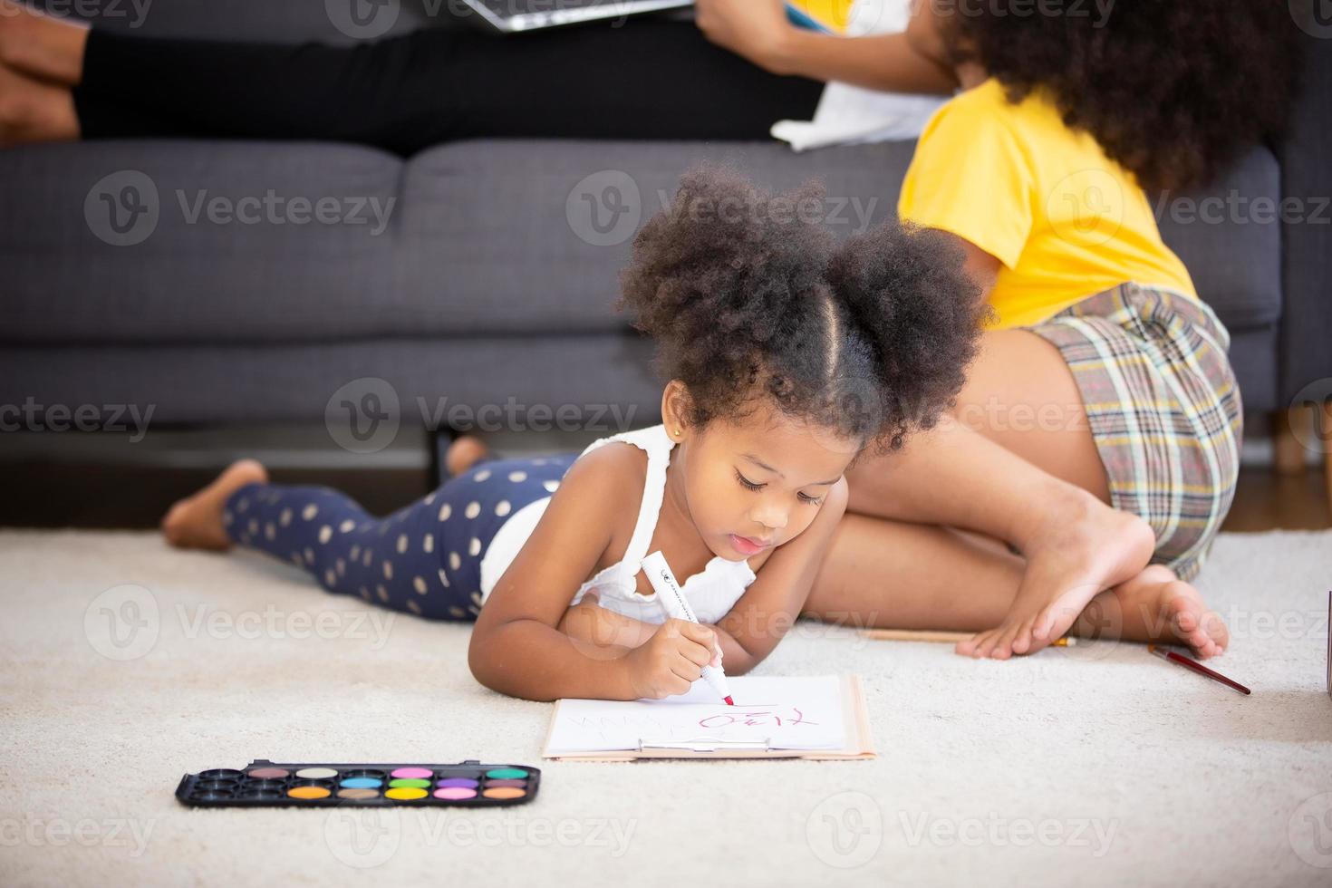 Happy black family spend free time together in living room at home. Couple sitting on sofa looks at little daughter show her drawing, son have a fun on warm wooden floor drawing with colorful pencils photo
