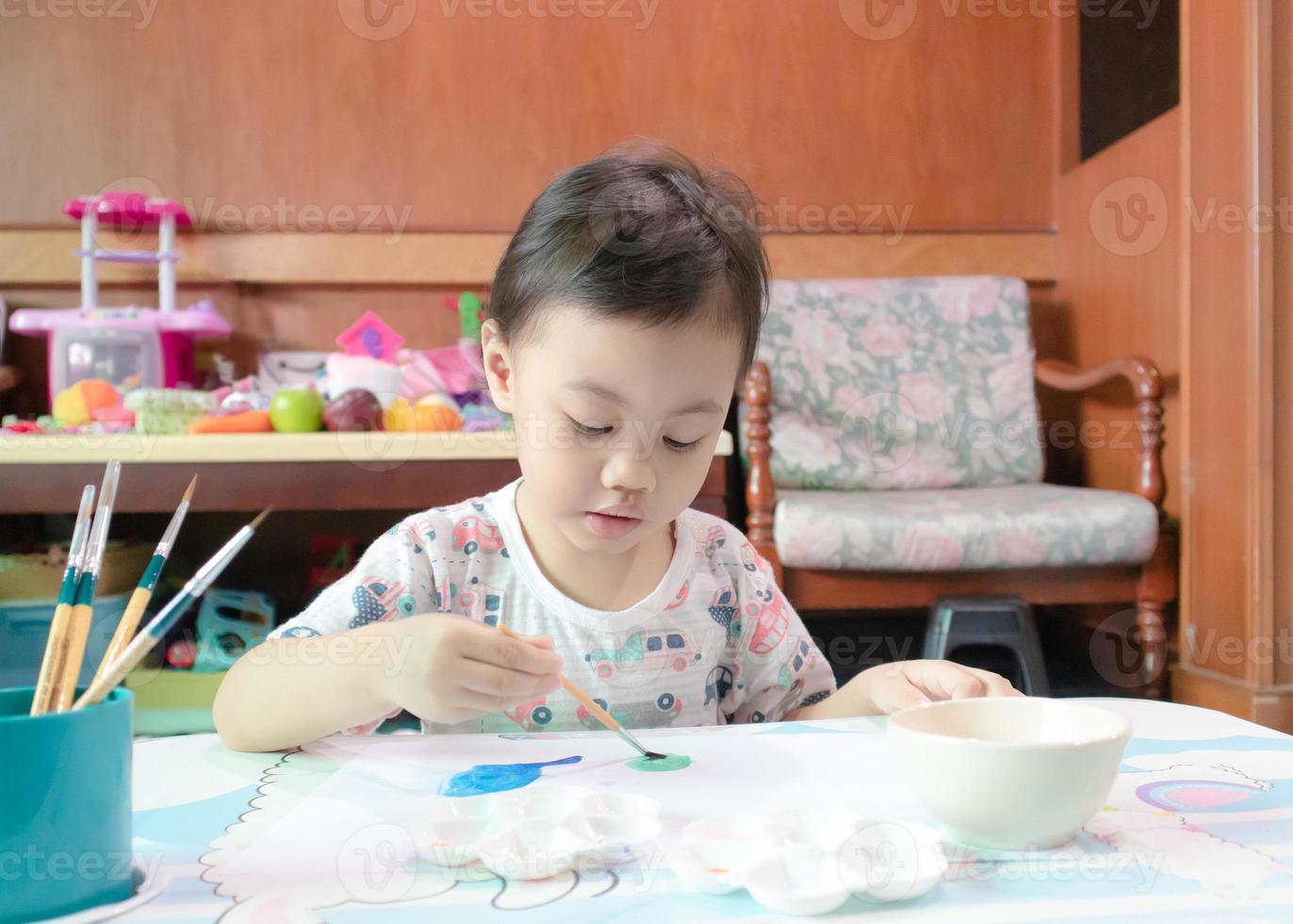 niña asiática, niña pequeña disfrutando de su pintura en papel blanco con pincel en casa. foto