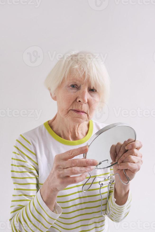 anciana triste mirándose en el espejo y tocándose la cara. edad, tiempo, concepto de cuidado de la piel foto