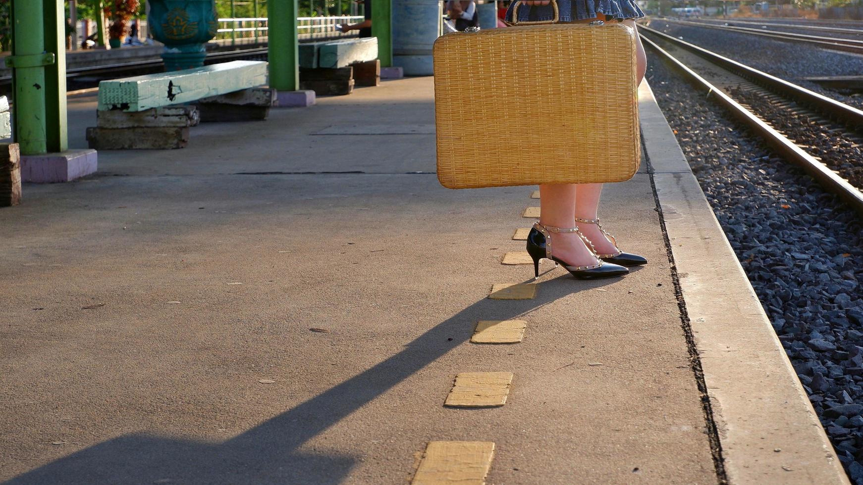 Low section of young female passenger holding vintage bamboo suitcase and waiting train with sunlight and shadow on platform at station in evening time photo