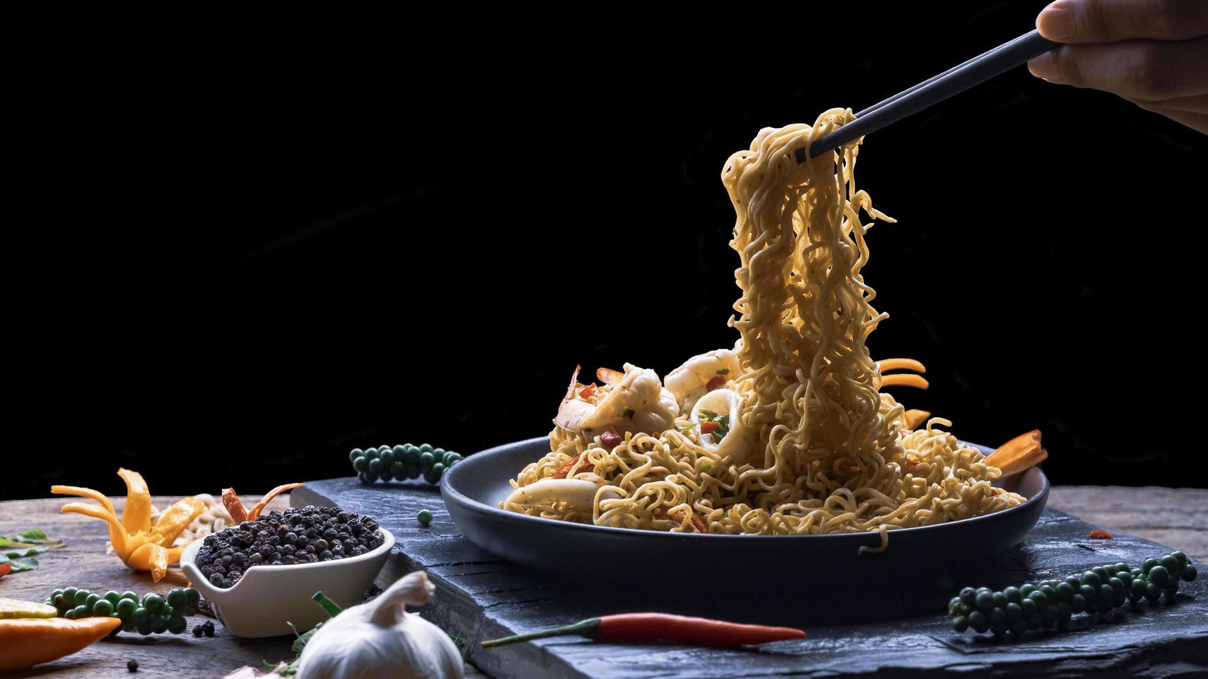 Man's hand using chopsticks to pick up stir fry instant noodles spicy seafood in black plate with various spices on black slate in black background, Asian meal concept photo