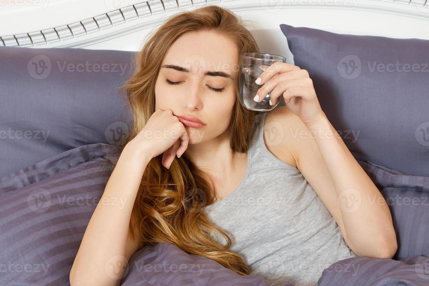 Happy young woman holding a glass of water and lying in bed in the bedroom - female hangover photo