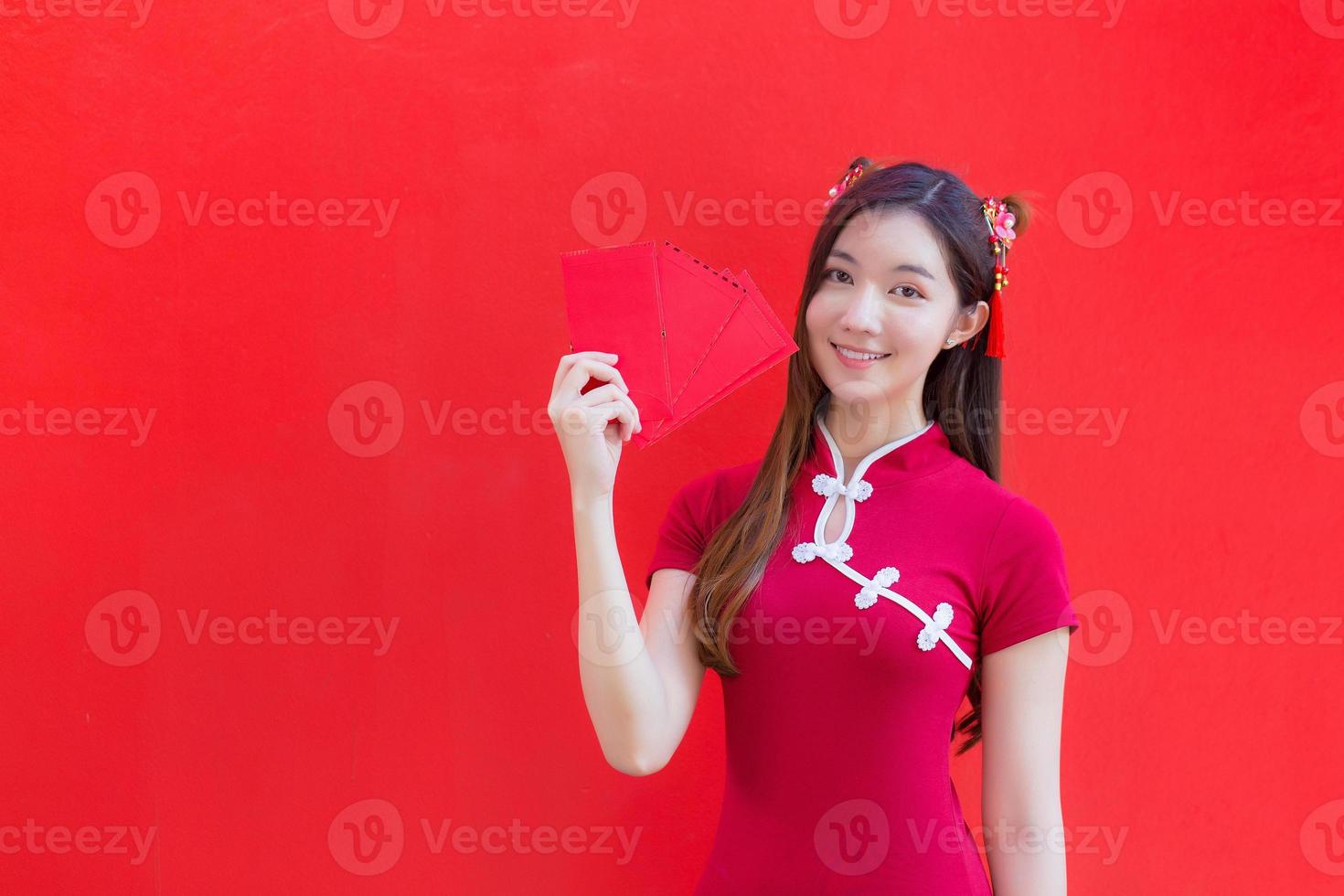 hermosa mujer asiática usa un cheongsam rojo y sostiene sobres rojos mientras mira a la cámara y sonríe alegremente con el fondo rojo. foto