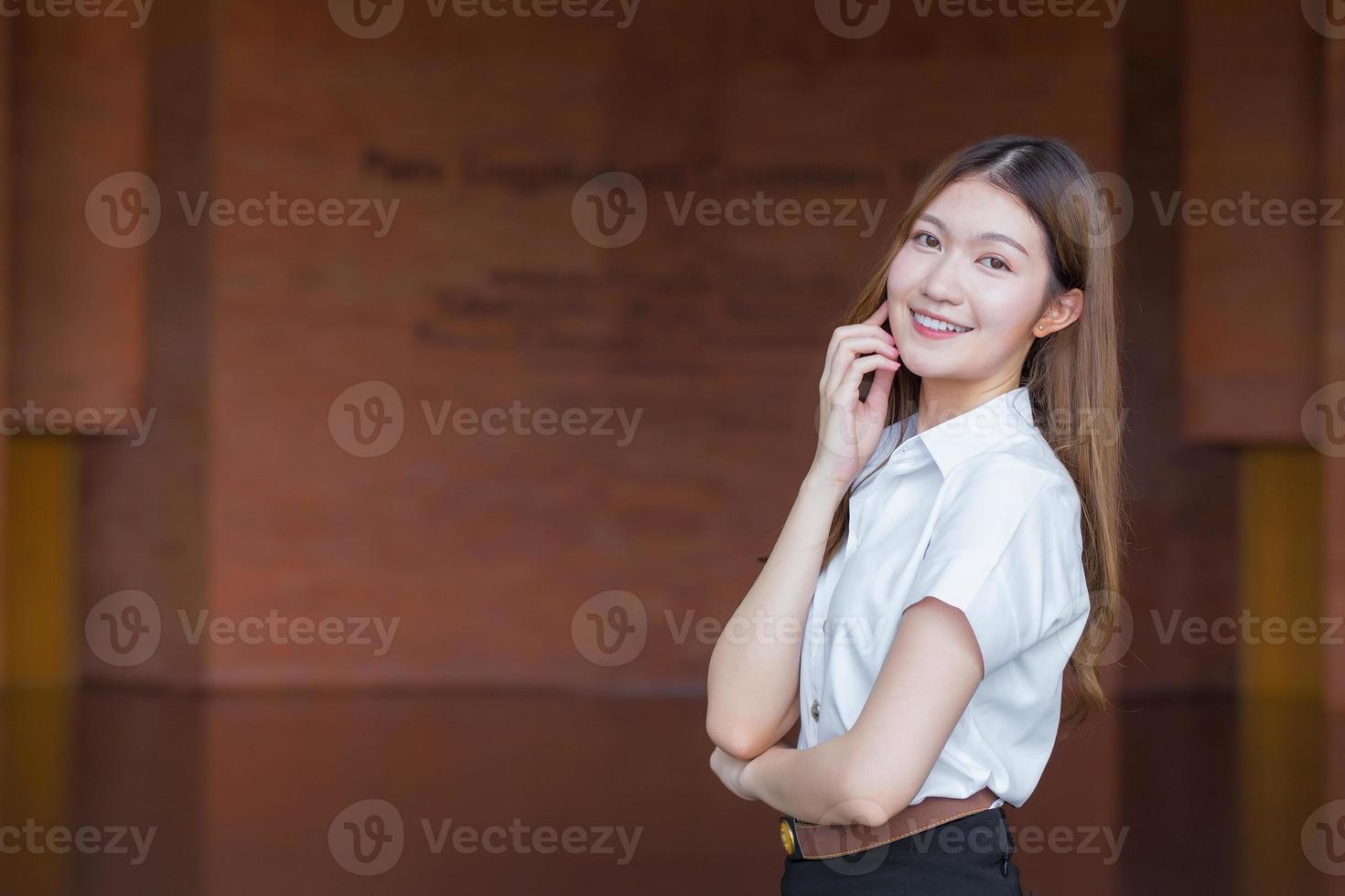 retrato de un estudiante tailandés adulto con uniforme de estudiante universitario. hermosa chica asiática de pie con los brazos cruzados sobre un fondo de ladrillo. foto