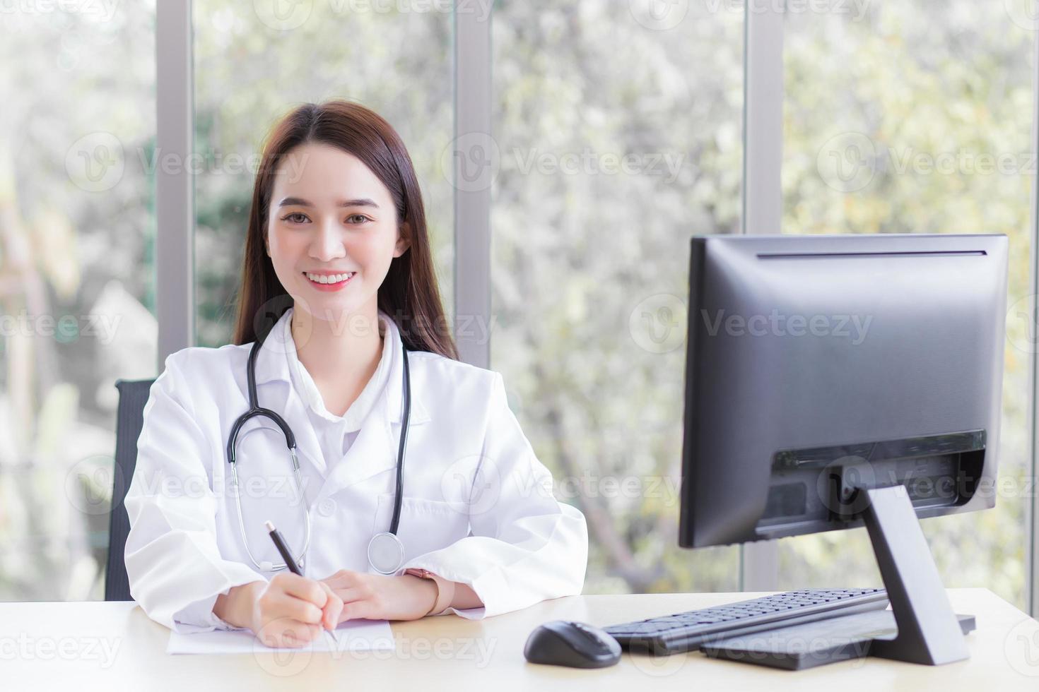 Professional Asian woman doctor wears medical coat and stethoscope while she is writing something on the paper and working in office room which she looking at the camera at hospital. photo