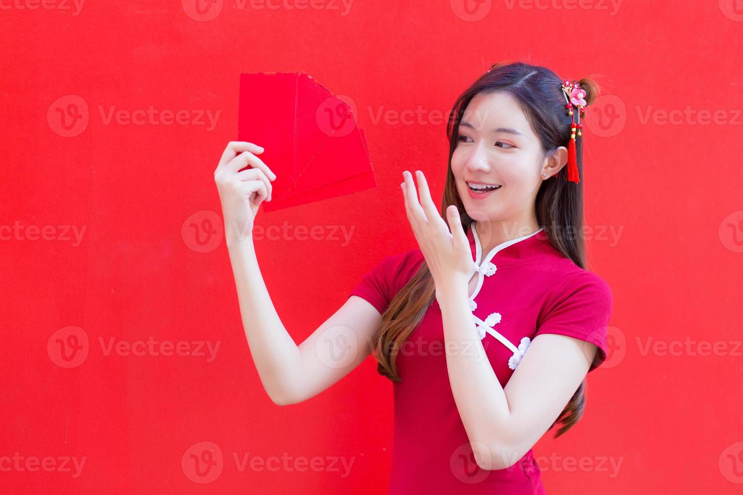 hermosa mujer asiática usa un cheongsam rojo y sostiene sobres rojos mientras mira a la cámara y sonríe alegremente con el fondo rojo. foto