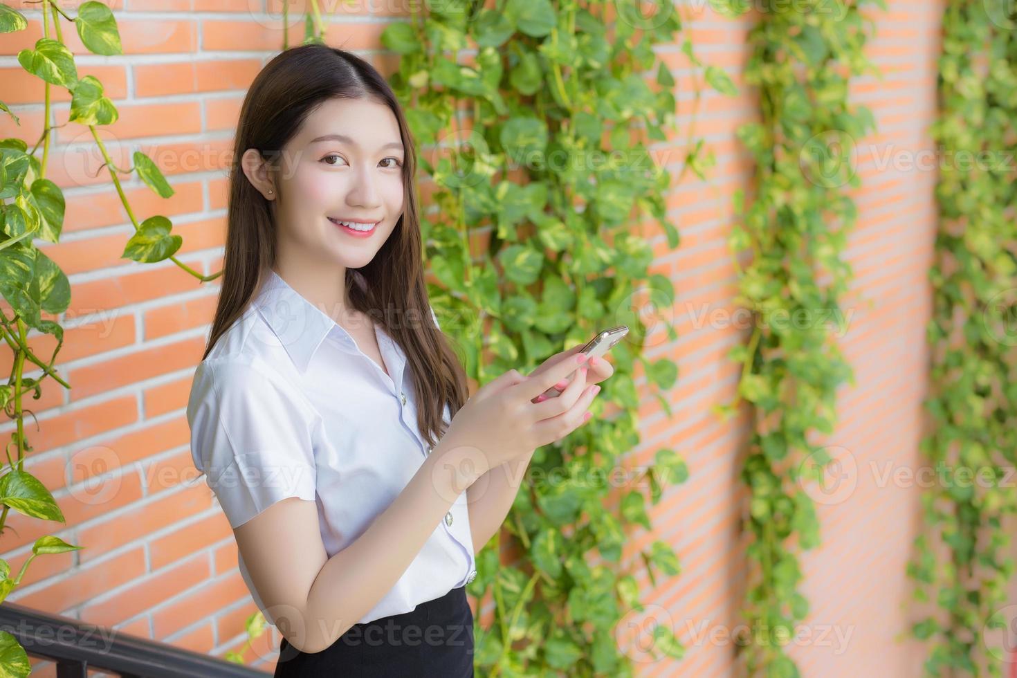 Portrait of an Asian Thai girl student in a uniform is smiling happily while using a smartphone at university with trees and walls as a background. photo