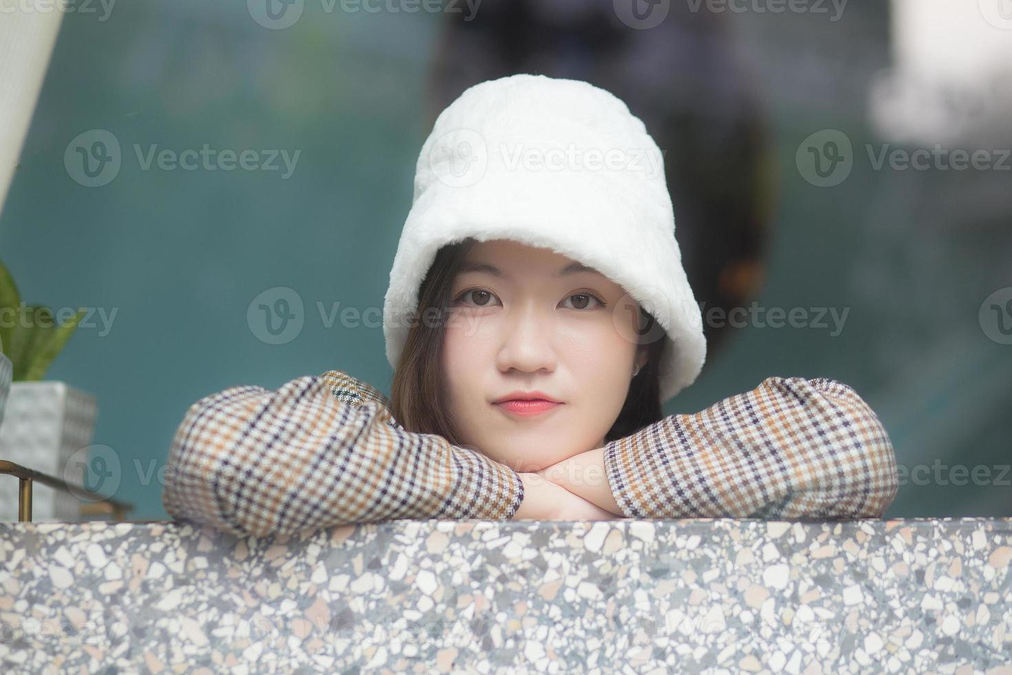 hermosa mujer asiática usa un sombrero blanco y un abrigo a cuadros mientras se sienta cerca de una ventana acristalada en el tema de año nuevo e invierno. foto