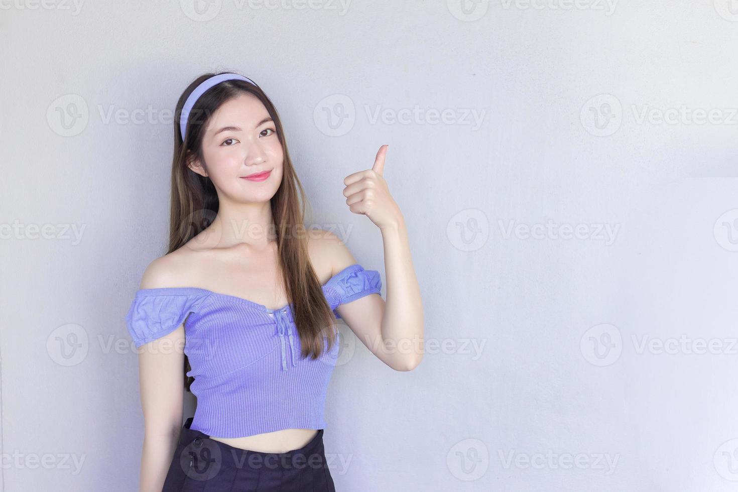 Young beautiful Asian woman wearing a purple shirt is acting thumb up as a good symbol and another hand shows as presenting something on the background. photo