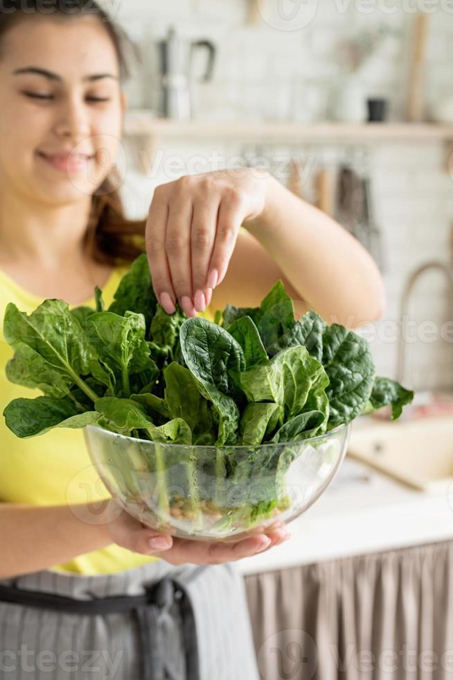 joven morenita mujer sonriente sosteniendo un plato de espinacas frescas en la cocina foto