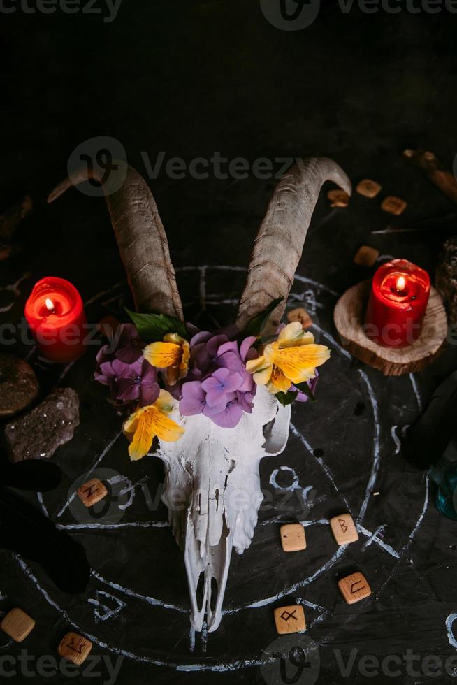 scull de cabra blanca con cuernos, flores, libro viejo abierto, velas en la mesa de brujas. foto