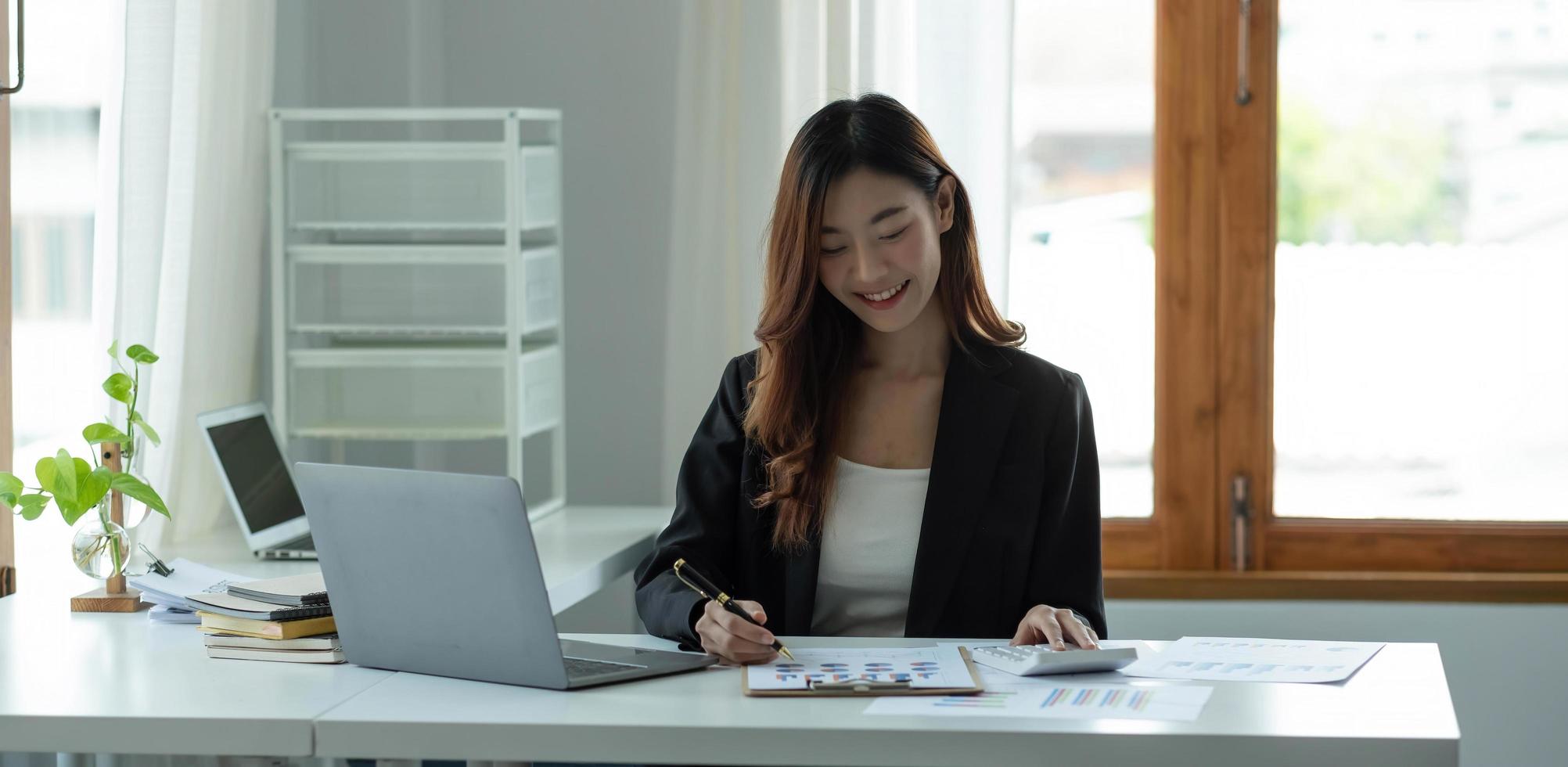 Beautiful Asian woman sitting in office using laptop and calculator. Happy business woman sitting at a desk in an office with a tablet computer. photo