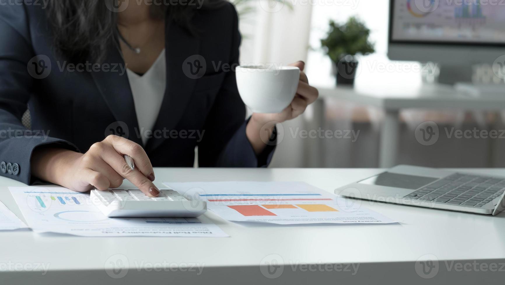 Asian businesswoman working on laptop computer and drinking coffee in modern office near window with calculator and business report on desk. work from home concept photo