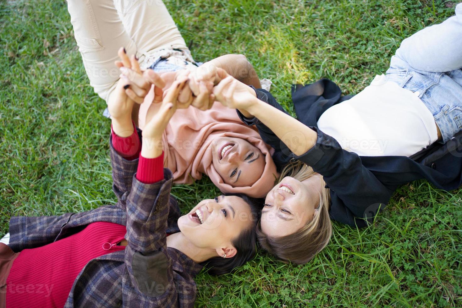 Joyful diverse women holding hands while lying on lawn photo