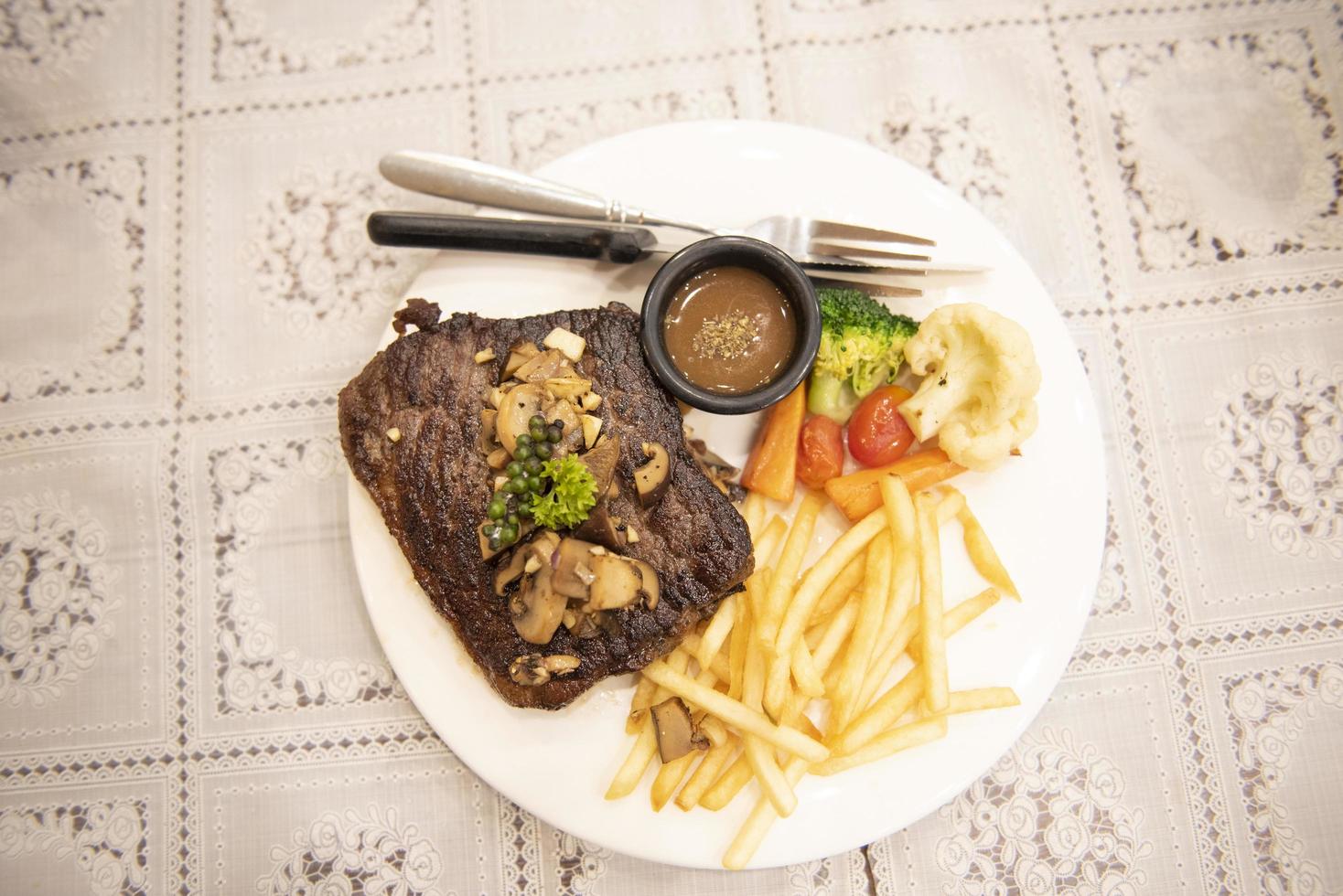 Bistec de carne asada con vegetales, hierbas, especias, pimienta negra, maíz y champiñones, bistec de ternera a la parrilla con papas fritas en un plato sobre la mesa del comedor. foto