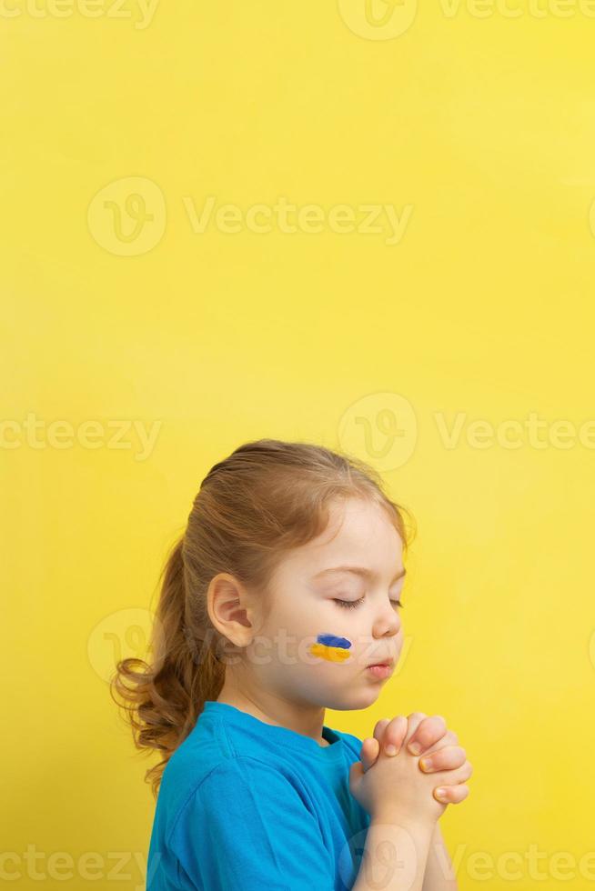 girl holding folded hands in prayer with yellow and blue colors of the Ukrainian flag on his cheek. Vertical photo