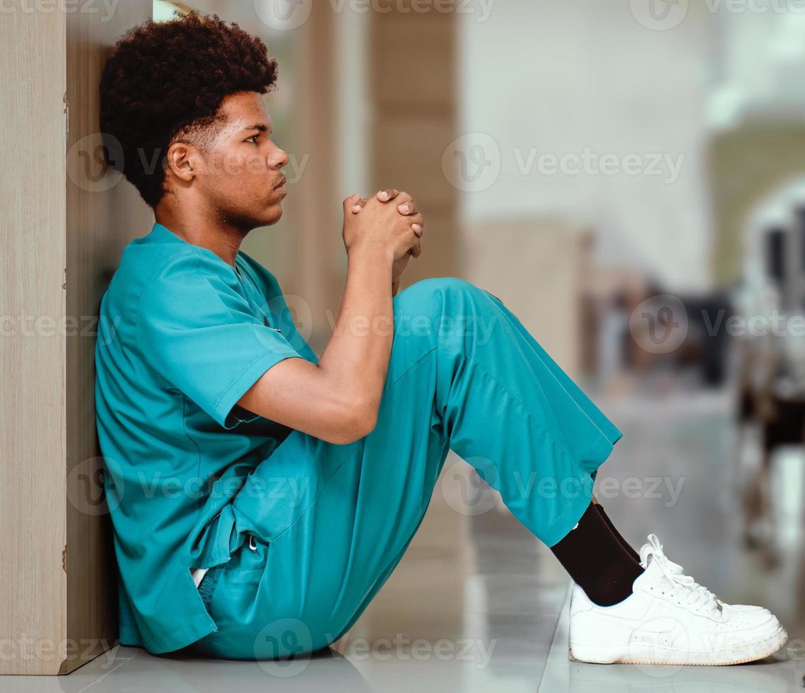 Emotional stressed intern doctor sitting against wall on floor in hospital corridor. Distraught medical student in seriously depressed, sad or exhausted. Overworked male nurse sits in despair or tired photo