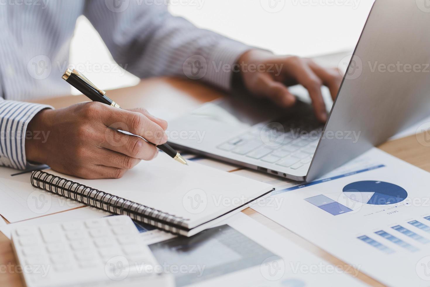 Businessman hand are taking notes on paper with a black pen, and using a laptop computer on a white desk in the office. photo