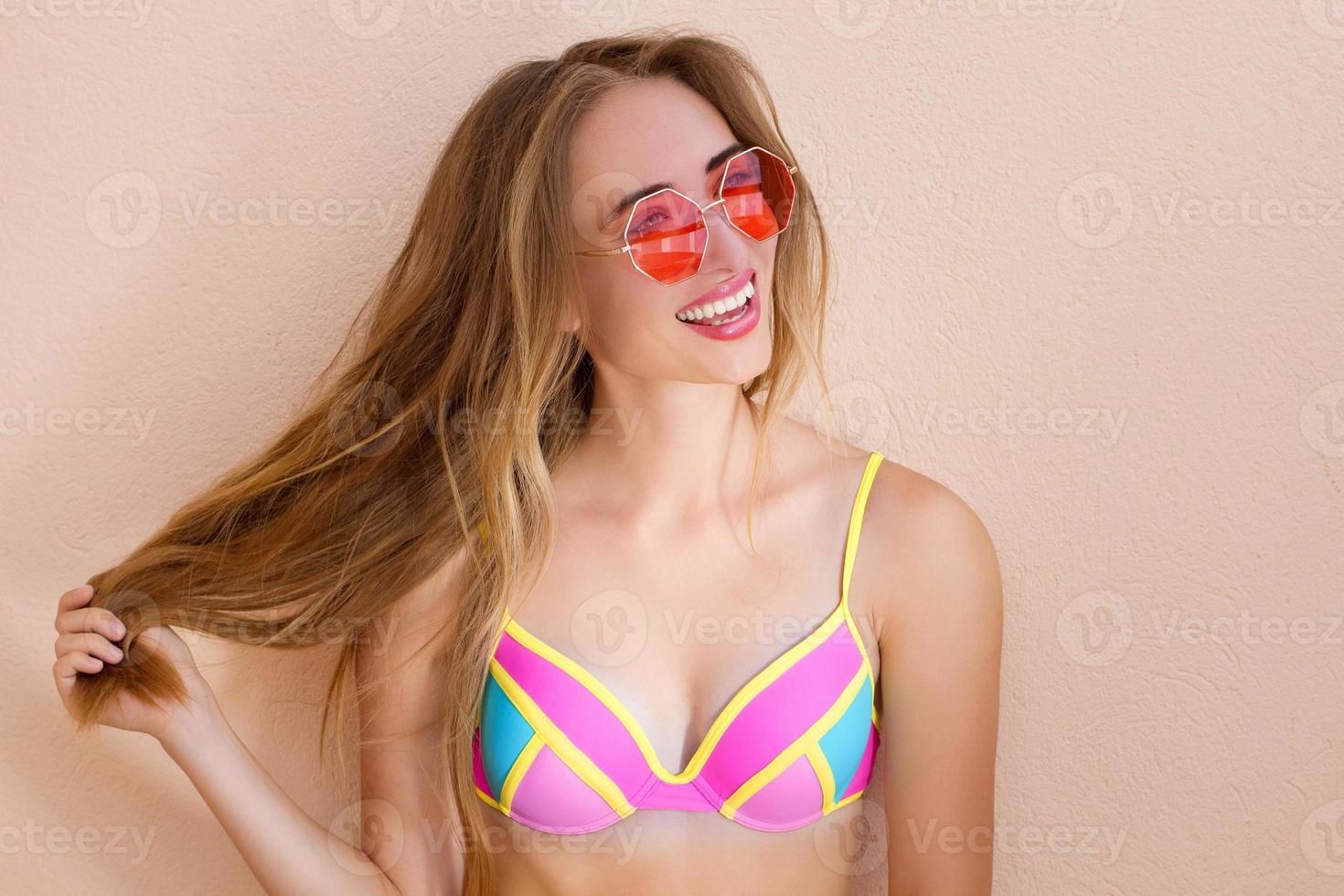 cerca de una chica feliz con gafas de sol rosas aisladas. vacaciones de verano y fin de semana divertido. concepto de verano. mujer joven sonriente en traje de baño de moda. enfoque selectivo. traje de playa de verano. copie el espacio foto