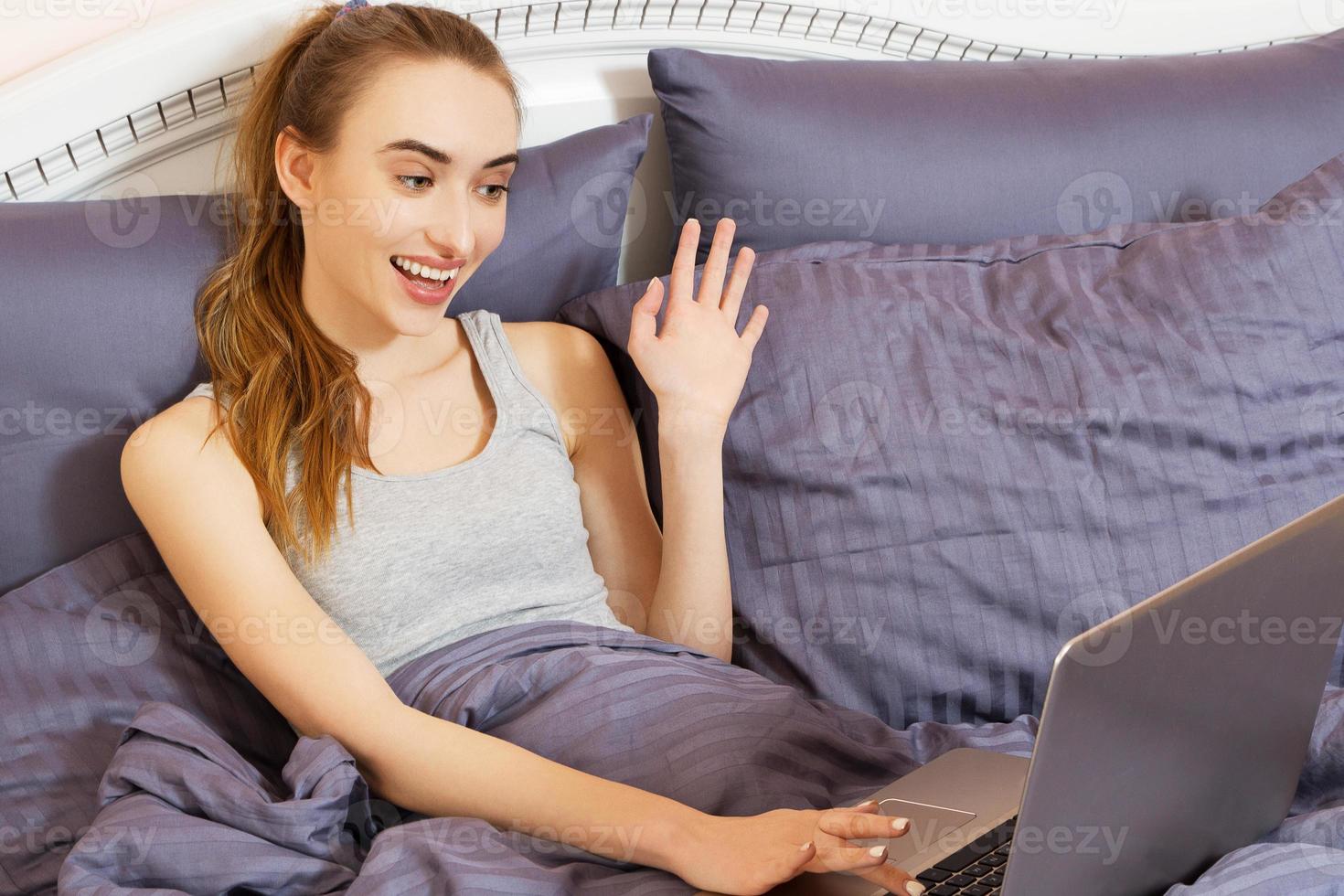 retrato hermosa joven blanca sonriente acostada en la cama relajándose usando el trabajo en su computadora portátil. rostro humano positivo expresión emociones concepto de satisfacción del servicio al cliente. foto