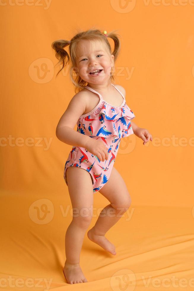 A little girl dressed in a swimsuit at the age of one and a half years is jumping or dancing. The girl is very happy. Picture taken in the studio on a yellow background. photo