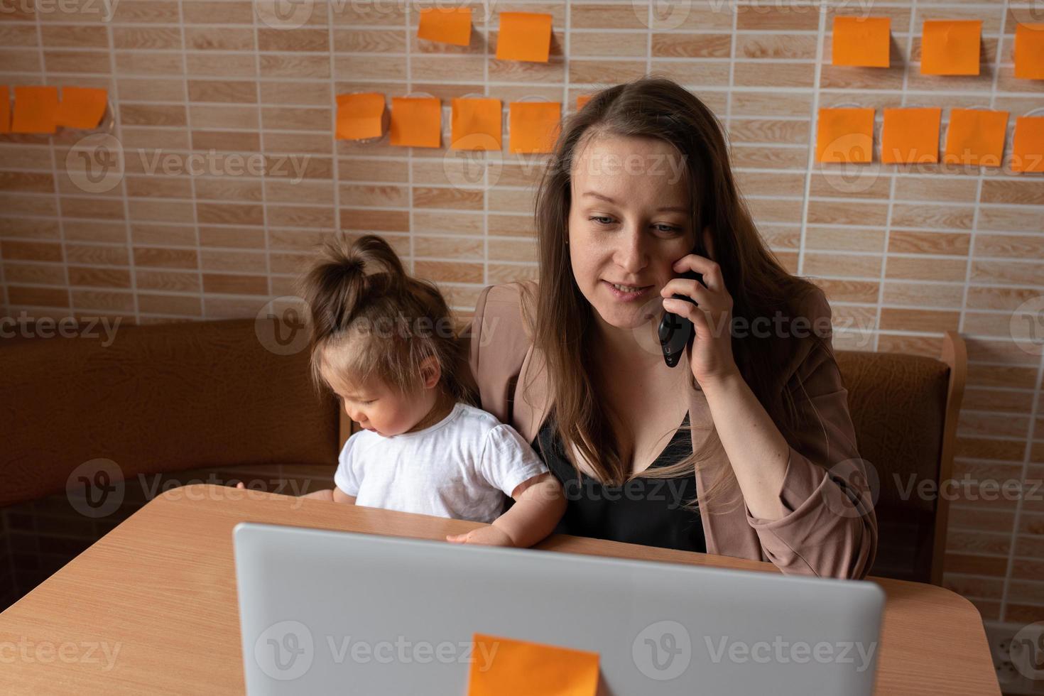 Young beautiful mother is trying to work remotely. Her little daughter is bothering her. photo