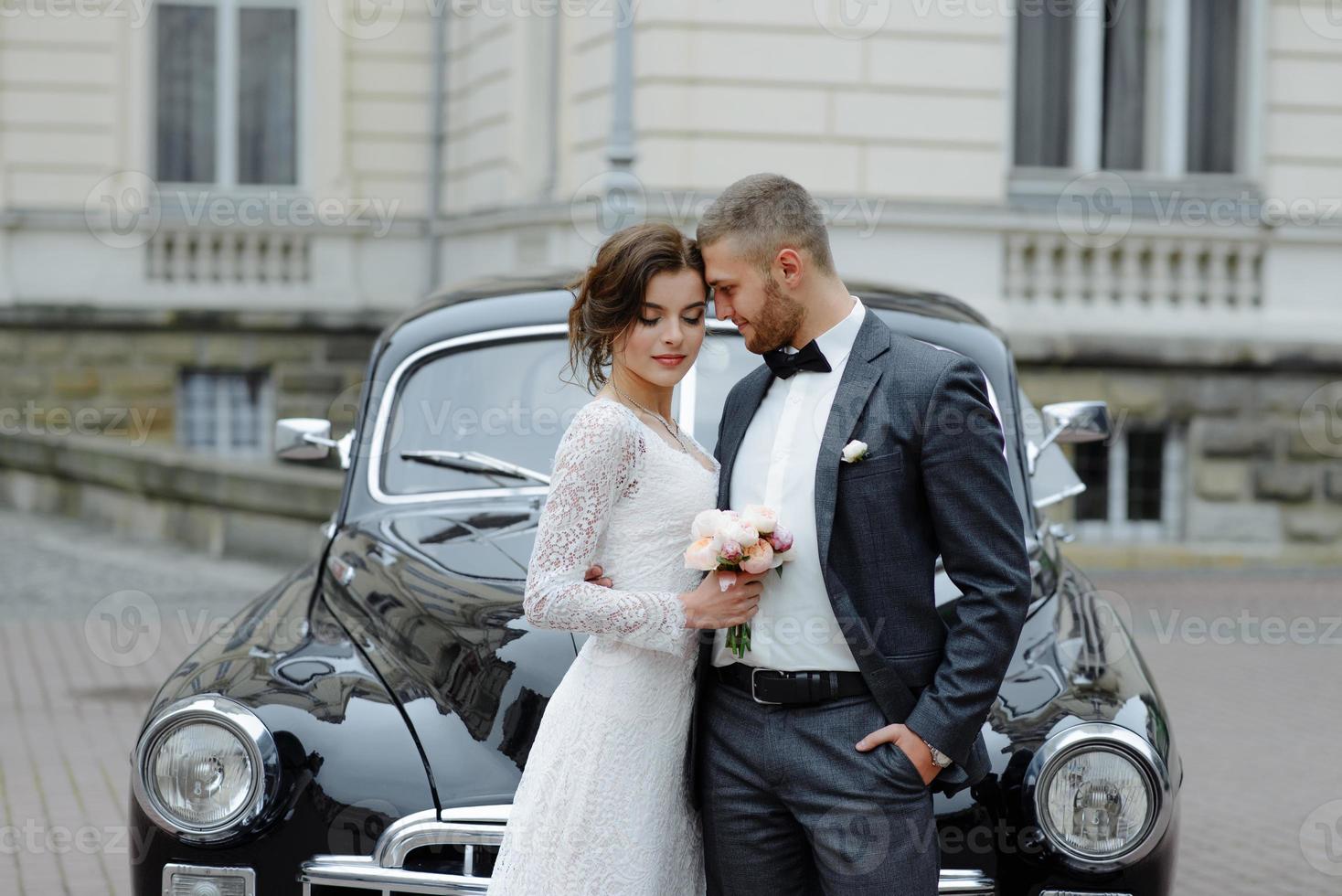 elegante novia hermosa y novio guapo abrazándose en un elegante coche negro a la luz. vista inusual desde atrás. pareja de bodas de lujo en estilo retro. foto