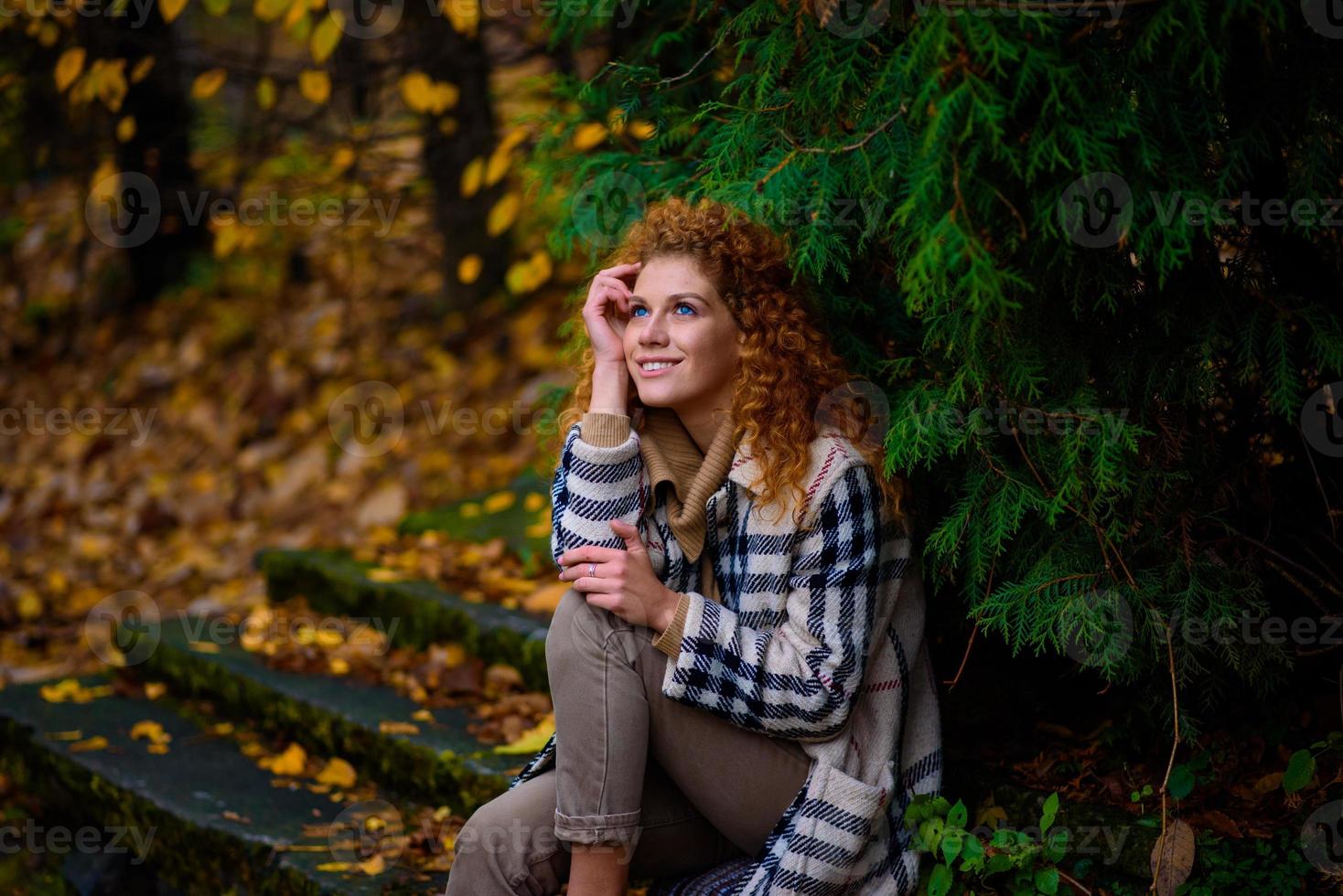 Beautiful red-haired girl with curly hair and blue eyes. The girl is wearing a checkered jacket. The girl is sitting on a bench. photo