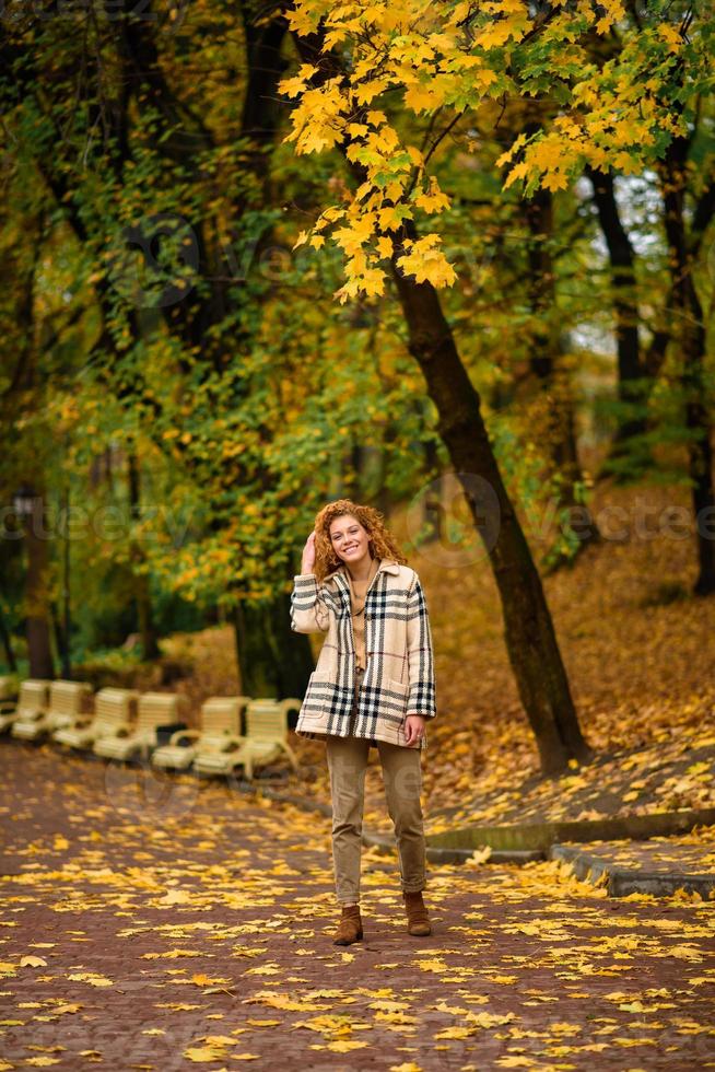 Beautiful red-haired girl with curly hair and blue eyes. The girl is wearing a checkered jacket. The girl is sitting on a bench. photo