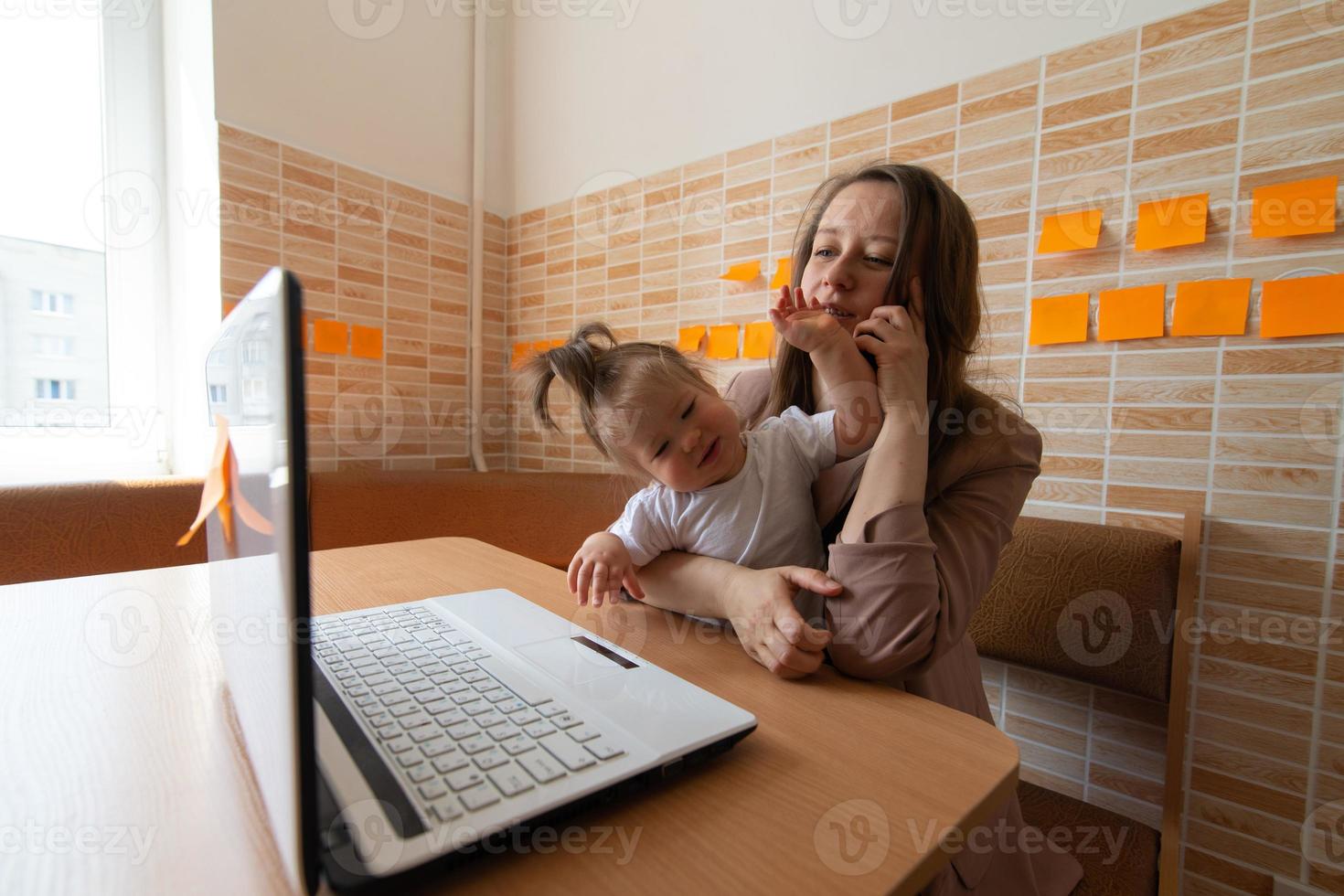 Young beautiful mother is trying to work remotely. Her little daughter is bothering her. photo