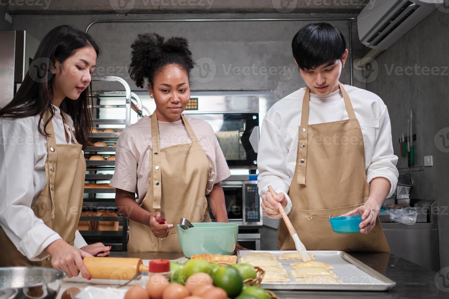 curso de cocina, un joven chef en delantal y un grupo de estudiantes de cocina, cepilla la masa de pastelería con crema de huevos, prepara ingredientes para alimentos de panadería, tartas de frutas en la cocina de acero inoxidable. foto