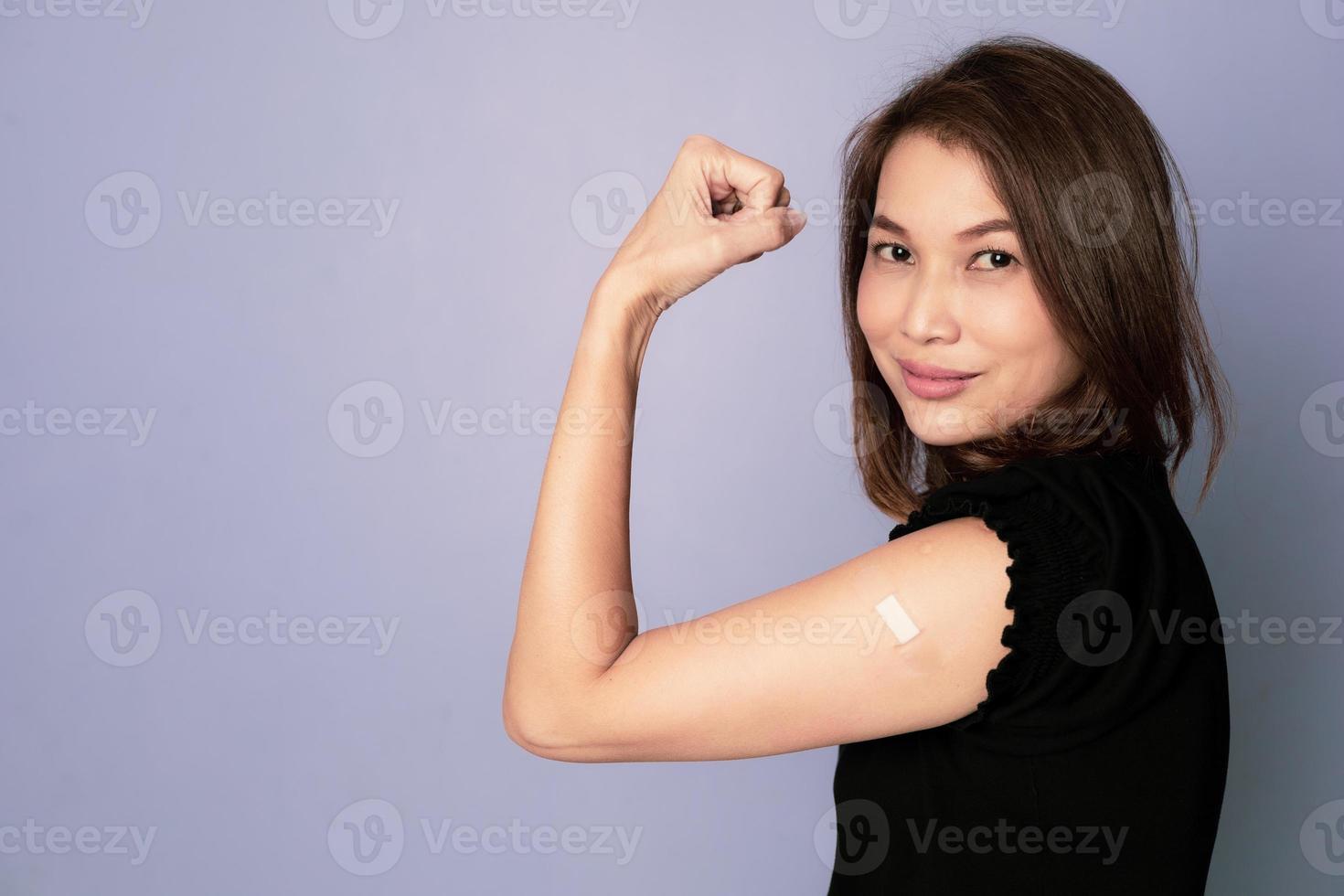 Portrait of vaccinated Asian woman showing plaster band aid on arm and show fist punch in winning and trust sign after coronavirus vaccine injection look at camera. Getting Vaccination Concept. photo