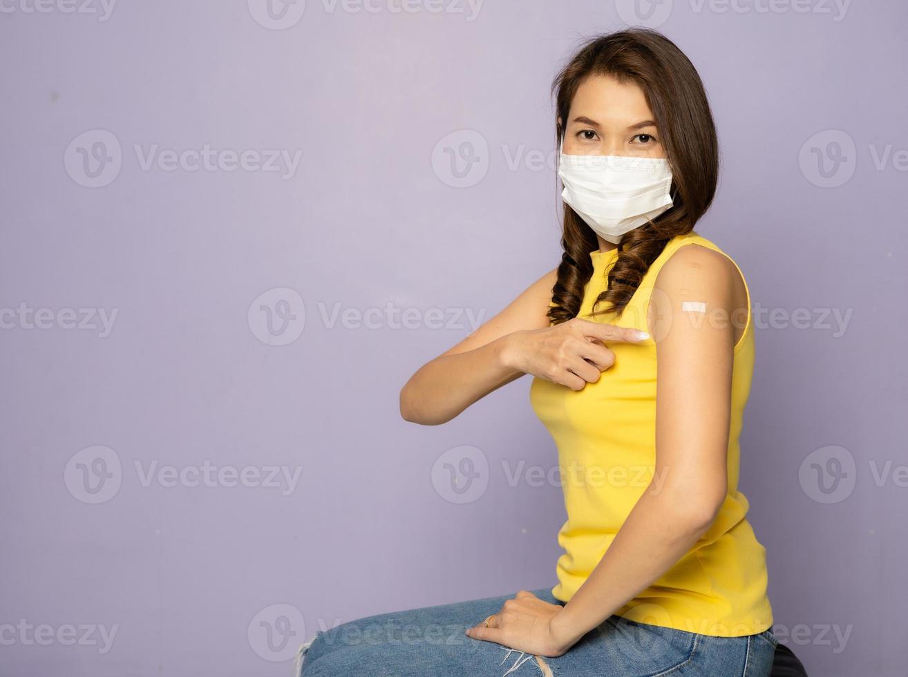Vaccinated Asian female showing plaster bandage on arm after getting Covid-19 vaccine injection while sitting wearing protective medical hygiene face mask. Coronavirus vaccination campaign concept photo