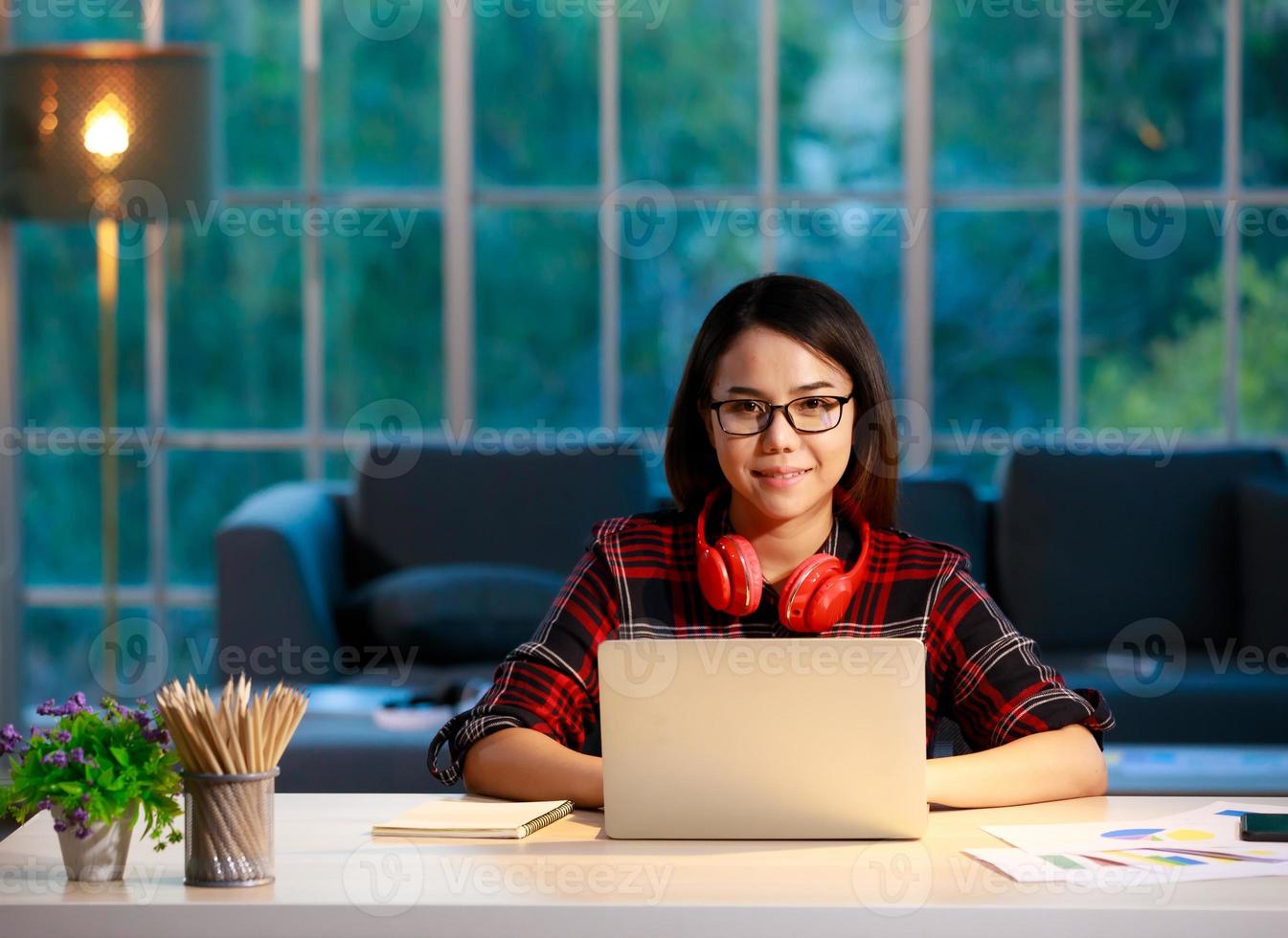 Cute Asian businesswoman working at home living room with twilight sky in outside of house background. She works with laptop notebook computer and looking to camera photo