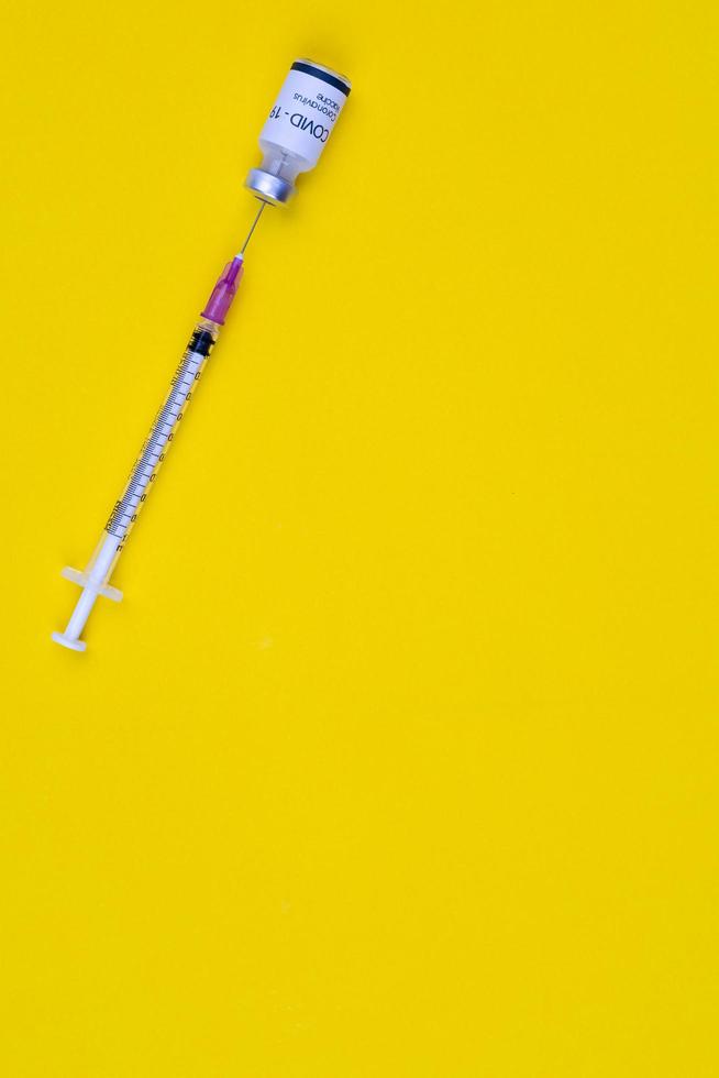Needle of syringe stab insert to bottle of Covid-19 coronavirus vaccine on yellow background with copy space. Top view shot photo