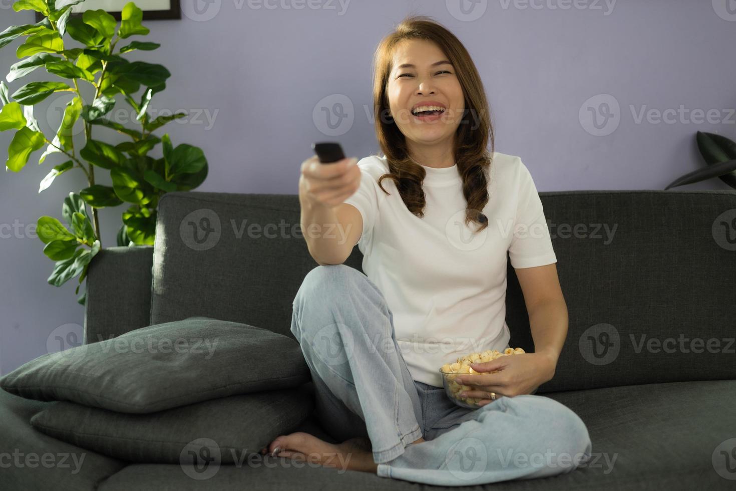 Cute Asian woman sitting on sofa with relax and cozy gesture holding remote control, watching television and laugh with fun and happy in home living room with sunlight from beside of her body photo