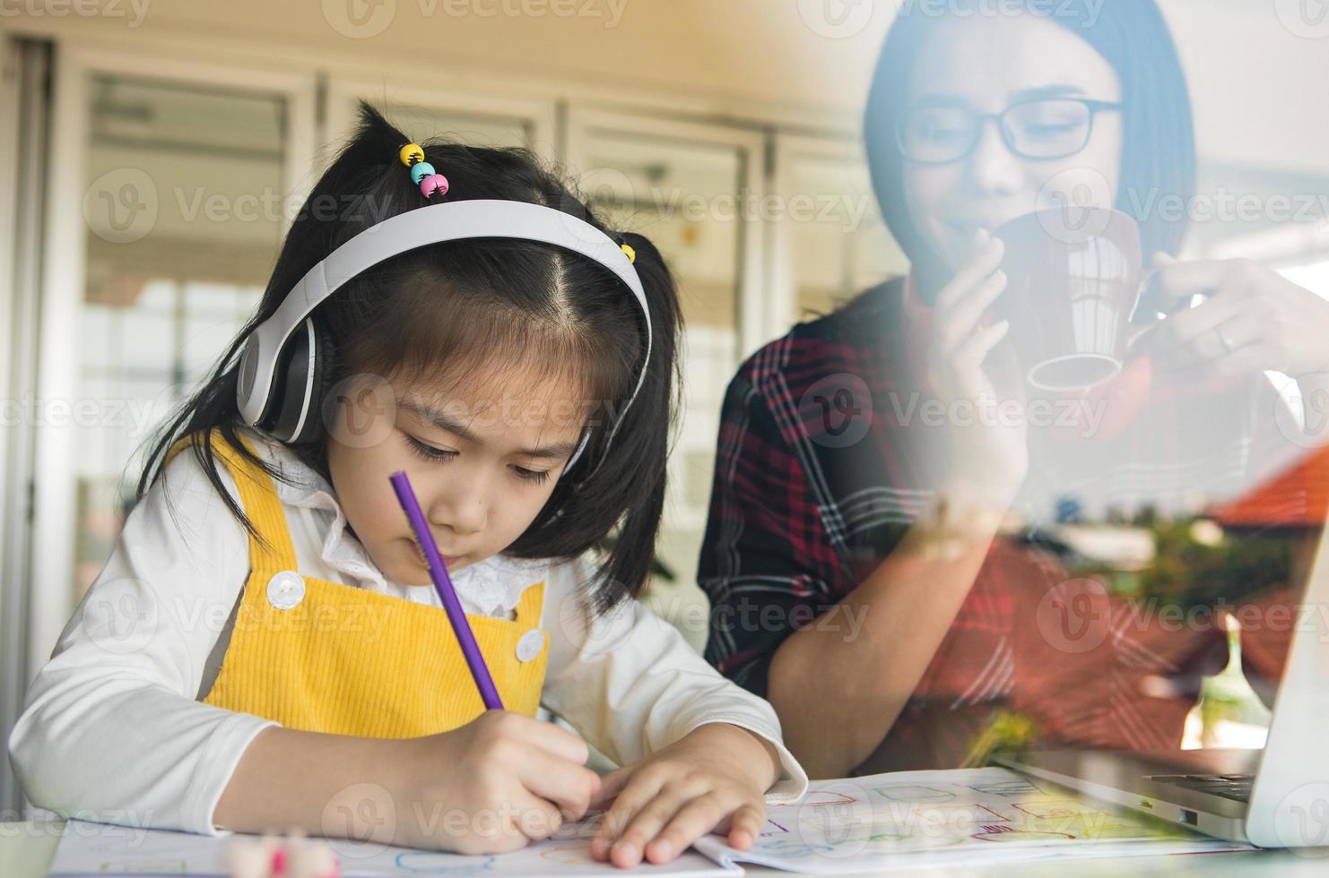 Asian mother holding coffee cup and and look at little daughter that doing homework beside her, She uses pencil to write on book photo