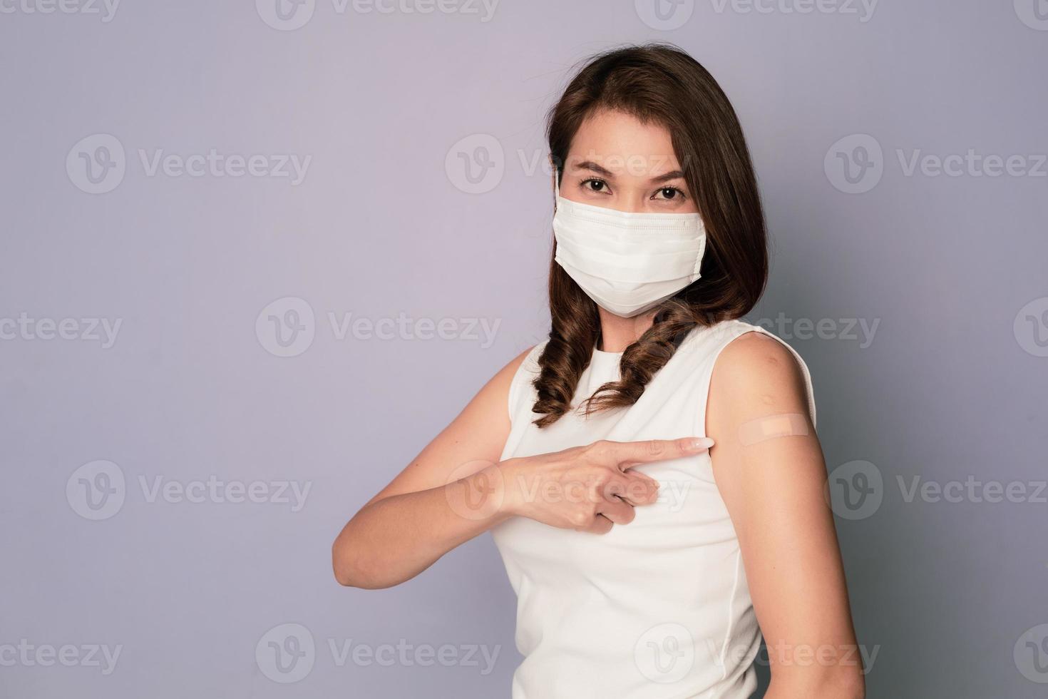 Vaccinated Asian female showing plaster bandage on arm after getting Covid-19 vaccine injection and smile while sitting wearing medical hygiene face mask. Coronavirus vaccination campaign concept photo