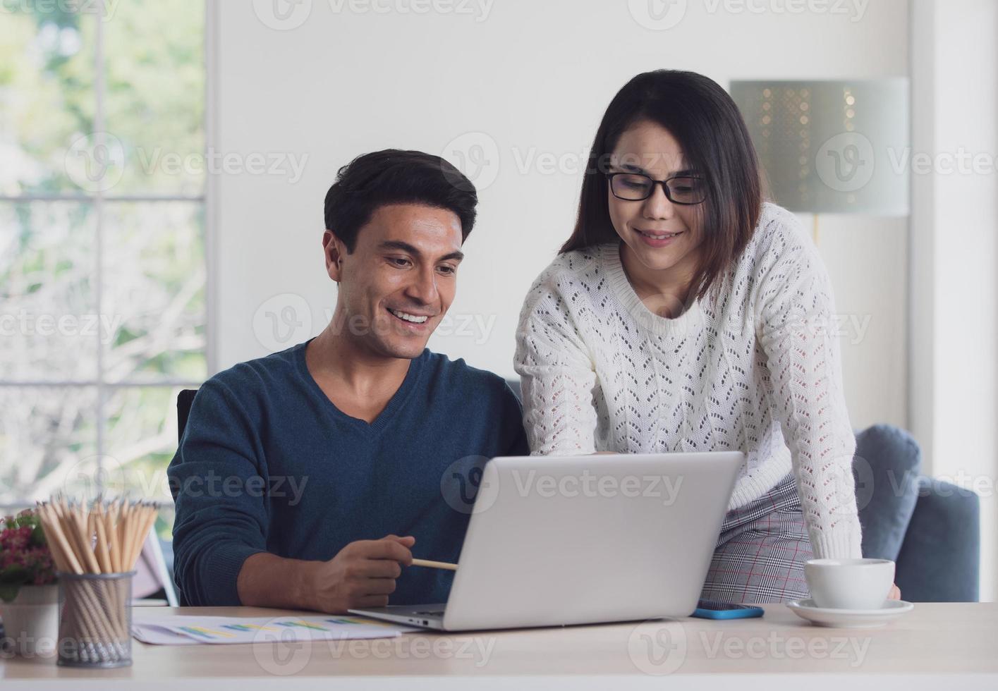 Mixed races couple family, Caucasian man and Asian woman, working together via laptop notebook computer and discussing in living room with happy and intimate. New normal work at home office concept photo
