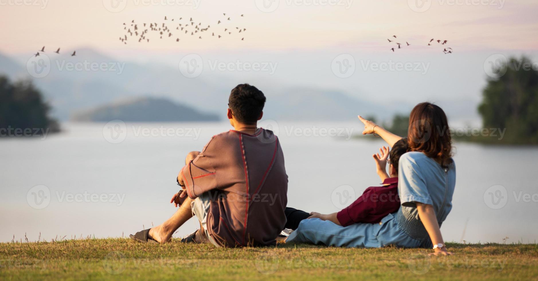 Mother and two sons sitting beside a big lake and see mountain view in the background, mom pointing finger to birds flying in sky. Idea for family tourist travels together photo