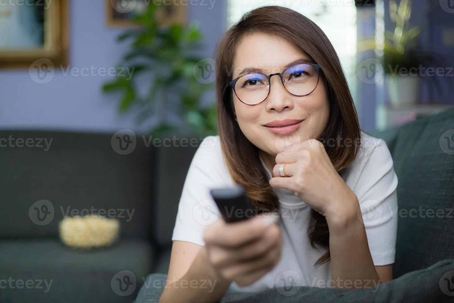 Cute Asian woman laying on sofa with relax and cozy gesture holding remote control aiming to tv, watching television and smile with fun and happy with popcorn blur in background. Hobby and relax idea photo