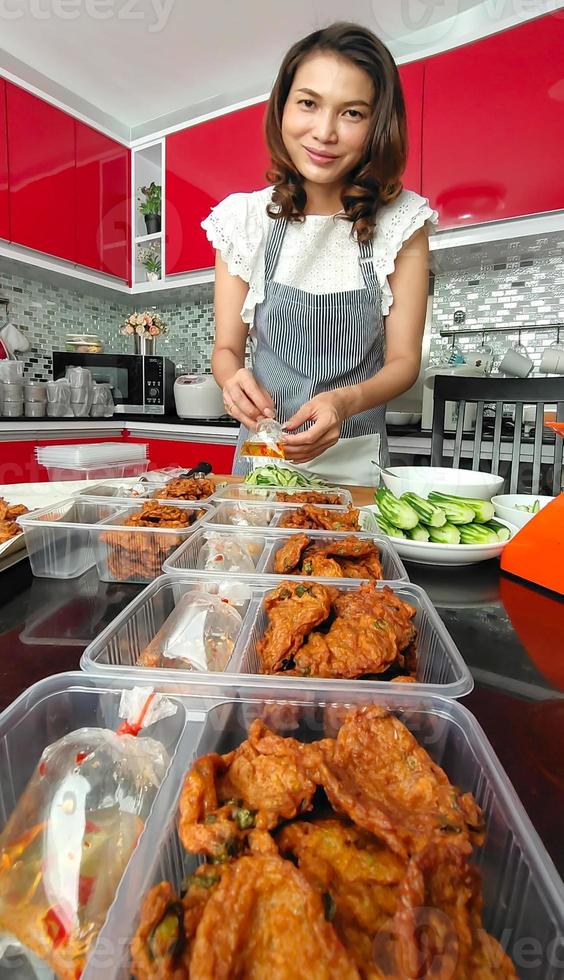 Beautiful Asian housewife and shop owner wearing apron preparing Thai style food,  fried fish-paste patty, for sale and ready to deliver for the customer. Idea for a side-hustle job at home photo