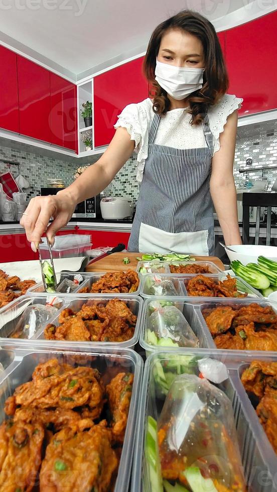 Asian housewife wearing hygiene mask preparing set of native Thai style food,  fried fish-paste patty, for sale ready to deliver for customers for extra income during coronavirus quarantine at home photo