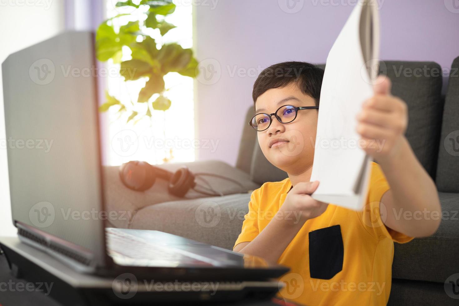 Young and cute 10s little Asian schoolboy wearing eyeglassed showing his homework for teacher during online studying by using laptop notebook computer in living room at home. Distance education idea photo
