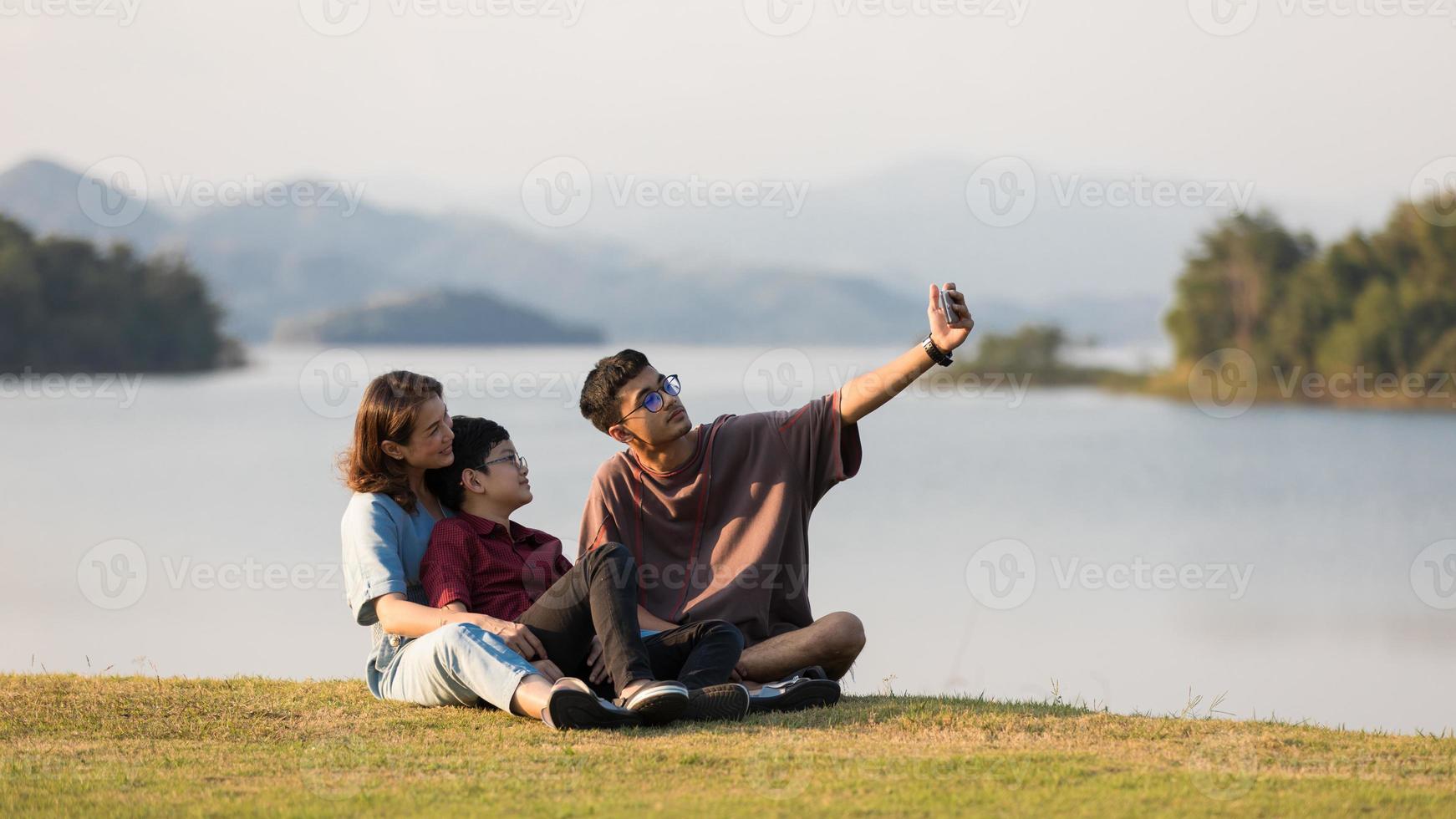 Asian family three members, mother and two young sons, sitting together beside huge lake with mountains and water in background. They using smartphone to take photos