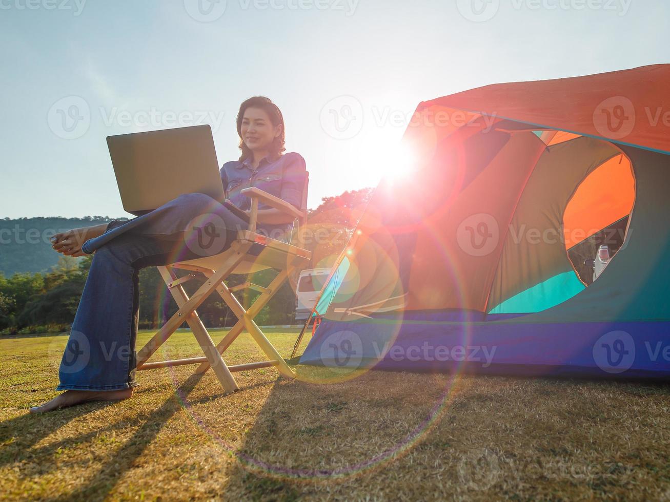 Beautiful middle-aged Asian woman sitting on wood chair in front of a colorful tent with circle light flare of sun rays of the morning in background. She uses laptop notebook computer to work outdoor. photo